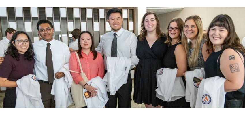 Seven students pose together holding white coats during an indoor medical school event.