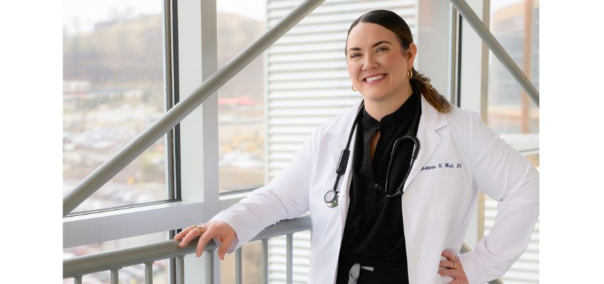 A smiling physician in a white coat with a stethoscope poses beside a large window.