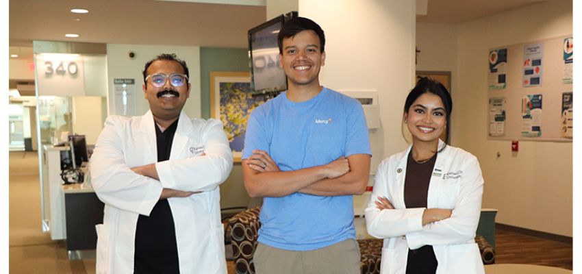 Two medical professionals in white coats pose with a man in a blue shirt inside a healthcare facility.