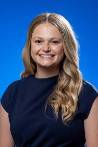 A smiling woman with long blonde hair poses for a professional headshot against a blue background.