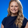 Asmilingwomanwithlongblondehairposesforaprofessionalheadshotagainstabluebackground