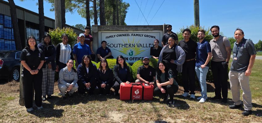 A group of 15 people pose outside the Southern Valley Fruit and Vegetable office with a red American Heart Association emergency response bag.