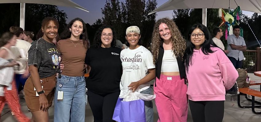 Six women pose together at an outdoor evening gathering, with several holding gift bags.