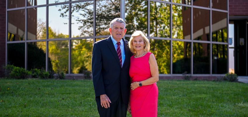A man in a dark suit and a woman in a bright pink dress pose together outside a building with large glass windows.