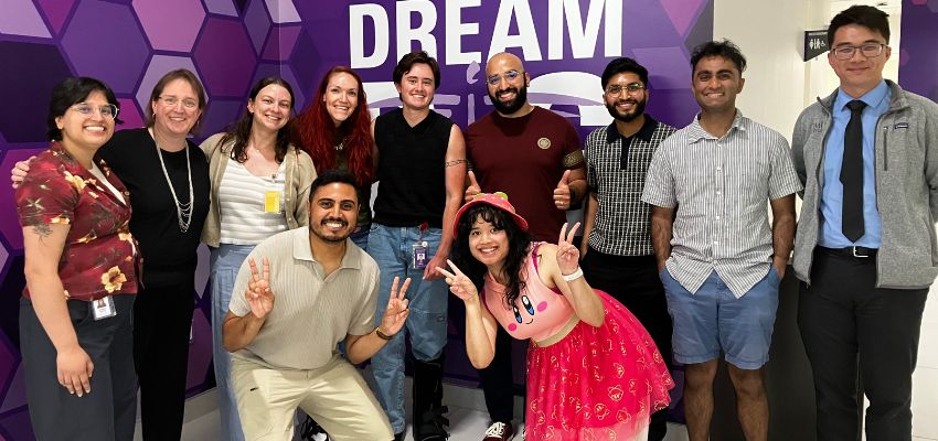 A group of 11 people pose together in front of a purple DREAM backdrop, with two participants crouching in front and making peace signs.