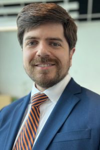 A smiling man with brown hair and a beard poses in a blue suit and striped tie for a professional headshot.