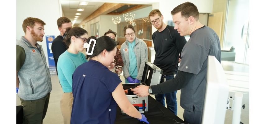 Students gather around a demonstration table as an instructor explains the use of medical equipment during a training session.