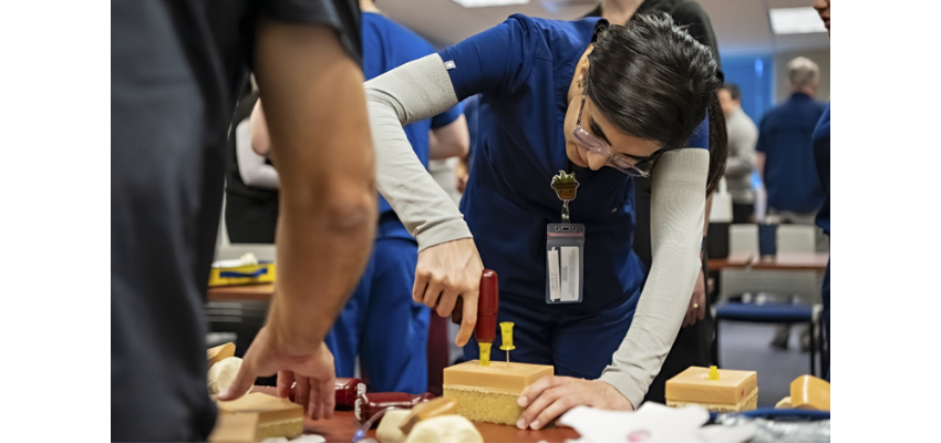 A medical student in blue scrubs practices inserting a needle into a simulation model during a hands-on clinical skills training session.