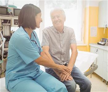 Doctor sitting next to elderly patient with her hands over his. Both are smiling.