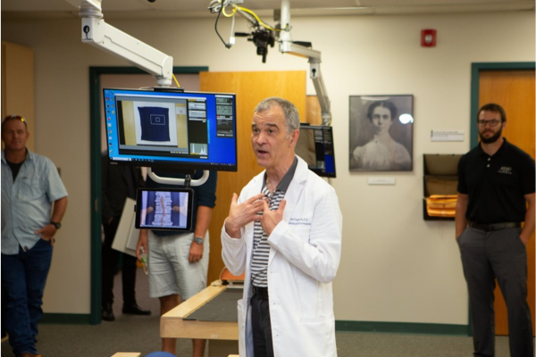 Dr. Degenhardt in the lab giving an OMM demonstration to visitors with a computer screen  next to him and spectators in the room