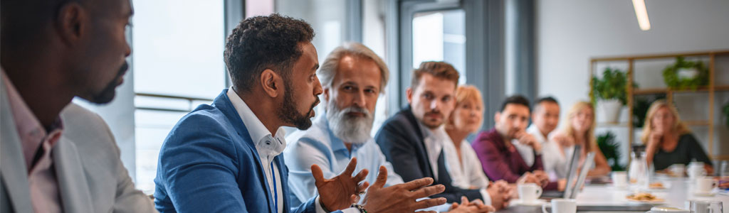 Black man in a suit speaking to a table of interested professional men an women