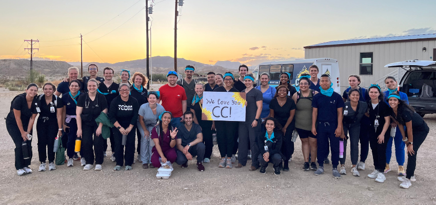 A group of medical students posing together outdoors at sunset, holding a sign that reads