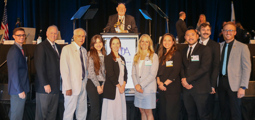 Medical professionals and students standing in a row at a formal event with a speaker at a podium in the background.